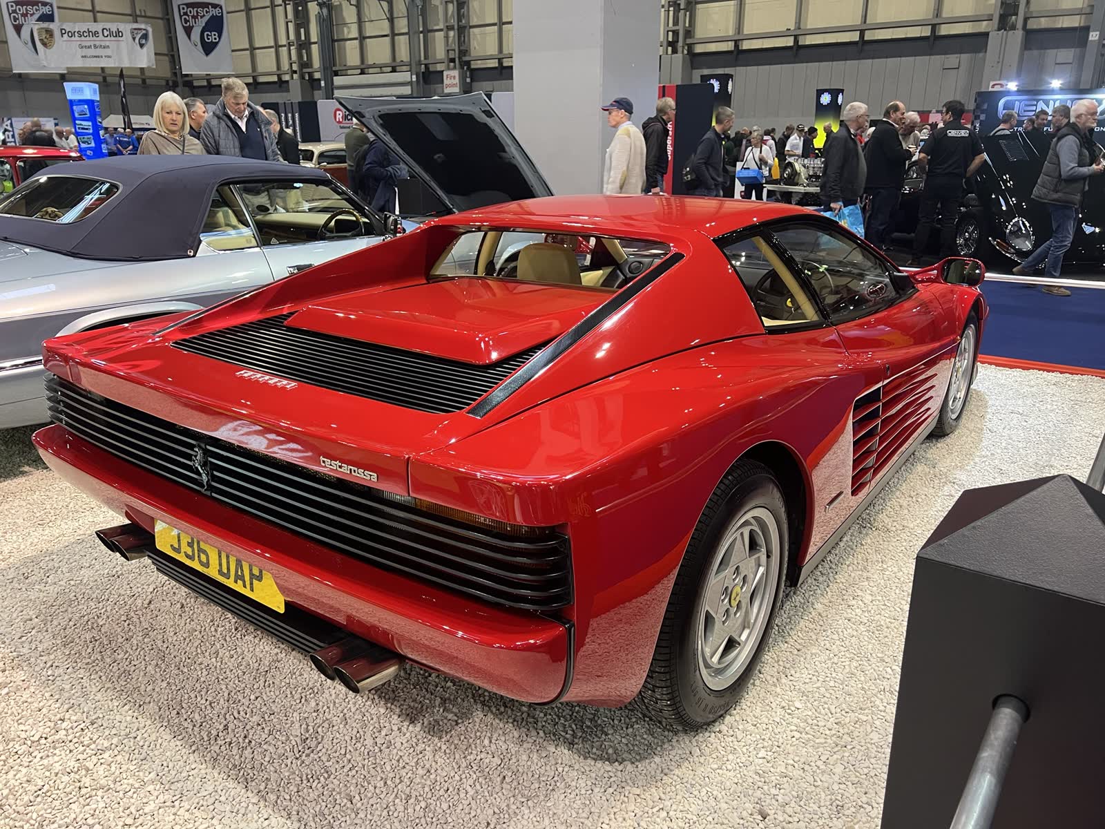 A red Ferrari Testarossa coupé at a classic car show, seen from the rear three-quarters, showing its fixed roof, slatted rear grille, quad exhausts and the side strakes along the doors