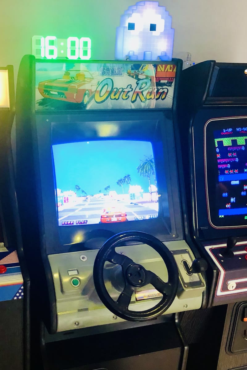 Alex's mini OutRun cabinet switched on in attract mode, with the OutRun marquee, the steering wheel without its centre badge and the Low/High gearstick, a Frogger cabinet beside it and a glowing Pac-Man ghost lamp on top