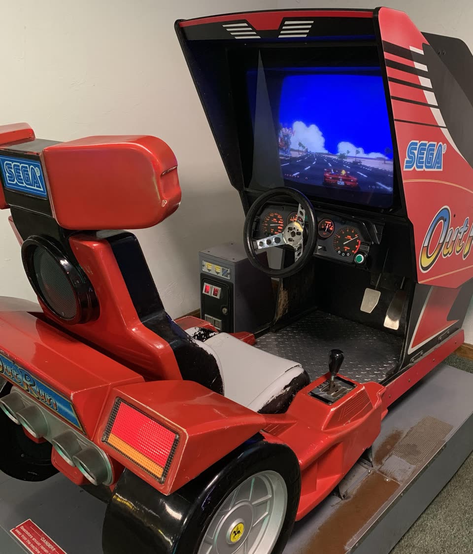 A Sega OutRun sit-down cabinet shaped like the back of a red Ferrari, with its moving seat and car body on a steel base, a steering wheel, dials and gearstick in front, and OutRun running on the monitor